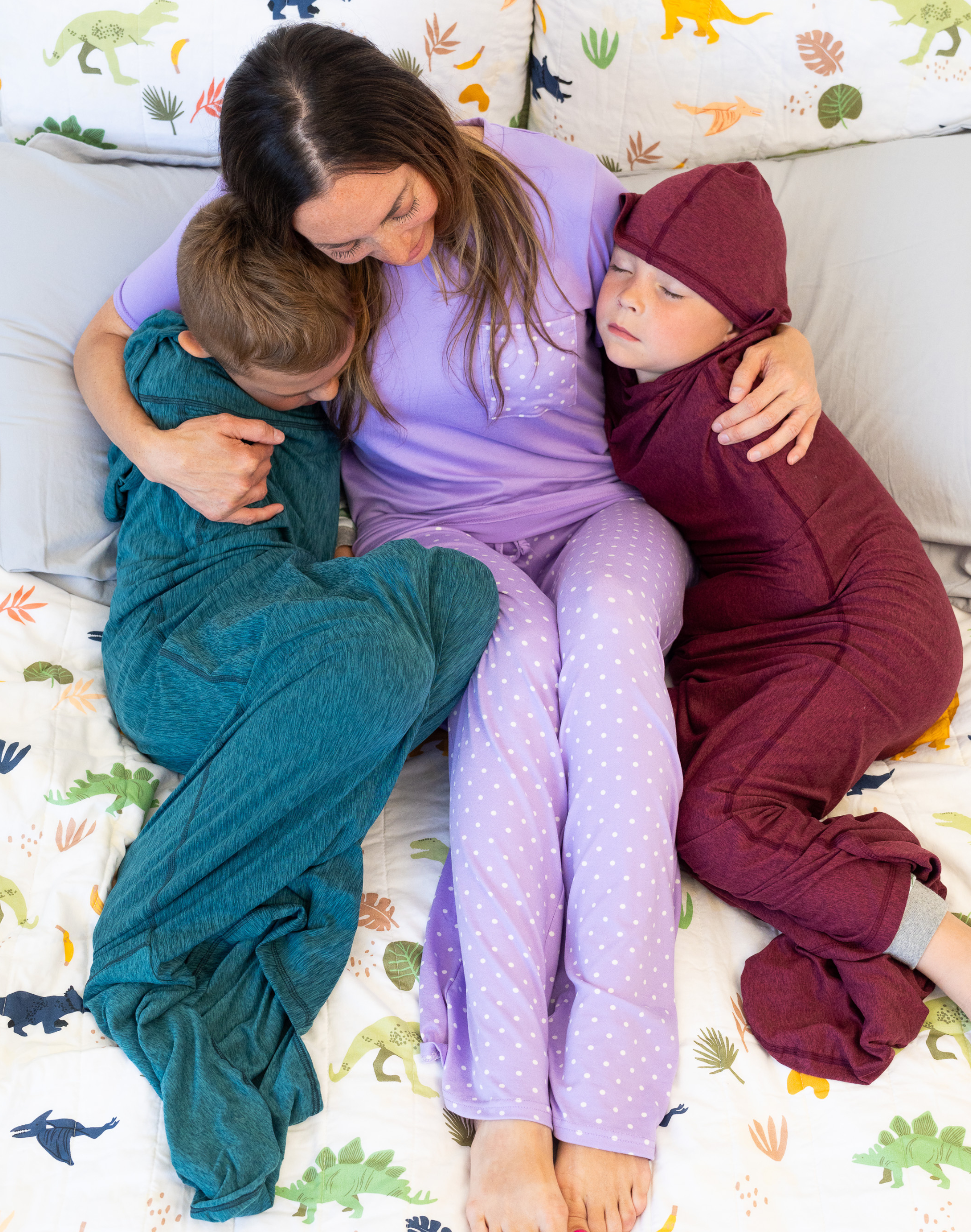 Woman sitting on a bed with two sleeping children, one in teal and the other in ruby, on a blanket with dinosaur patterns.