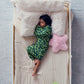 Child resting on a cream bed sheet while wearing a green camo Junior Sleep Pod with white pillows and a pink star pillow nearby.