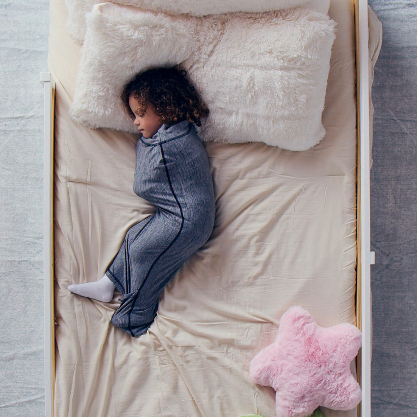 Child sleeping on a cream bed sheet while wearing a navy ribbed Junior Sleep Pod with a pink star pillow nearby.