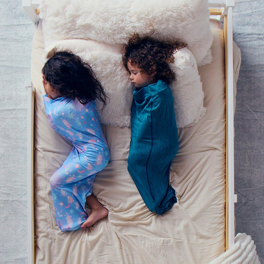 two-children-resting-on-a-cream-bed-sheet-while-wearing-turquoise-and-cosmic-blue-star-pattern-junior-sleep-pods-under-white-pillows