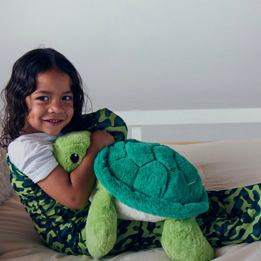 child-smiling-in-bed-while-wearing-a-green-camo-junior-sleep-pod-and-hugging-a-plush-turtle