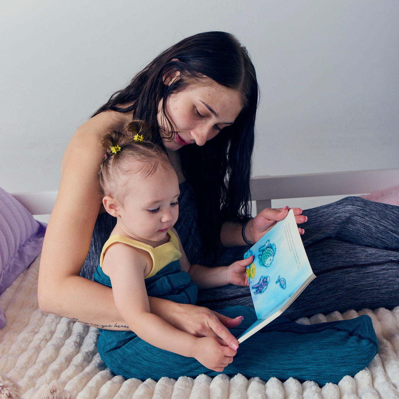 Adult reading with a young child on a bed while both are wrapped in turquoise and graphite color Sleep Pods.