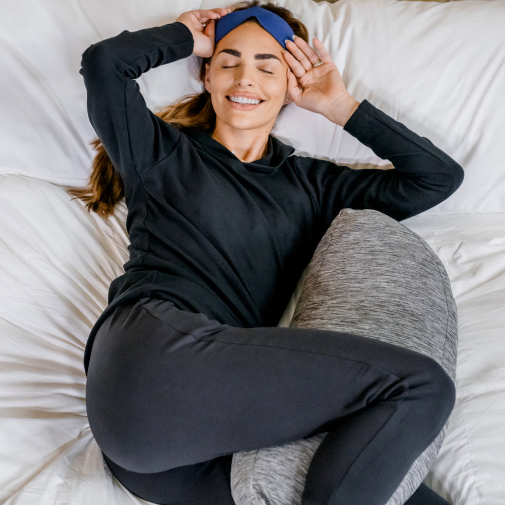 woman holding  Pod Pillow™ between knees laying in bed waking up with sleep mask on