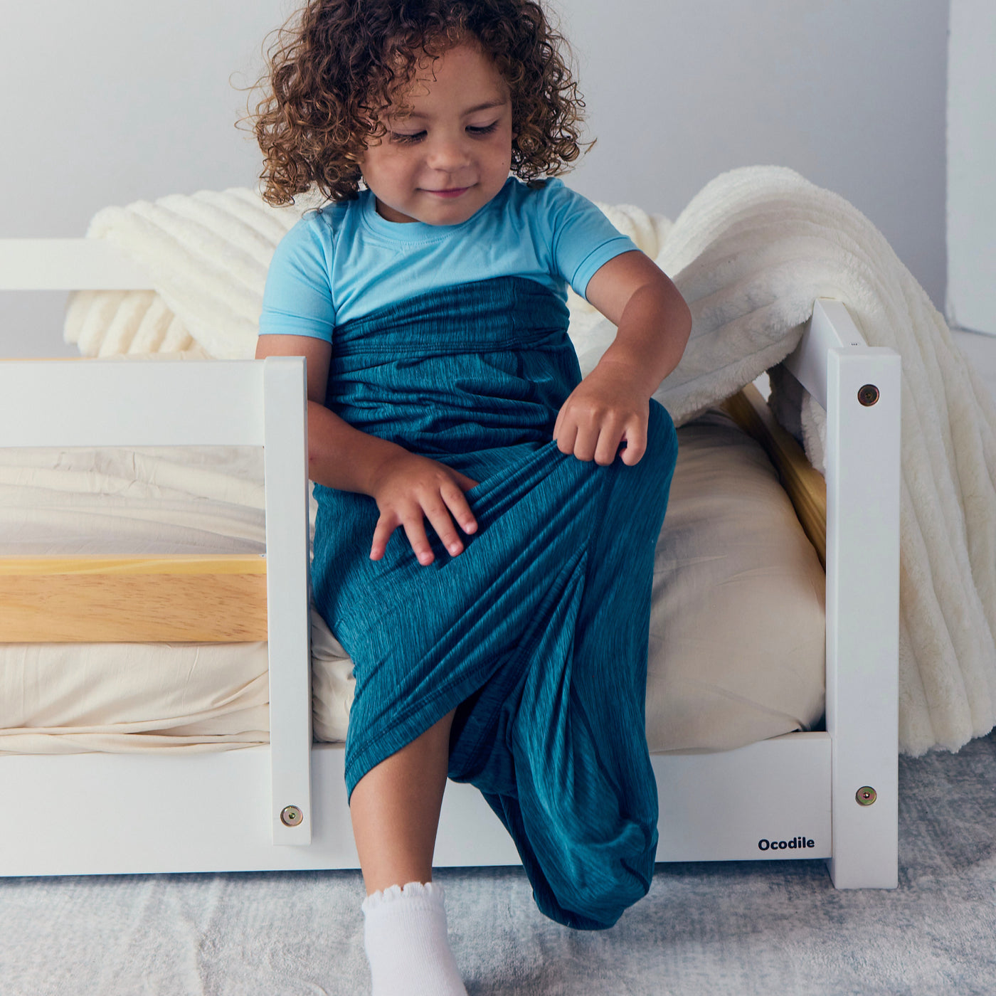 Child sitting on a low bed while wearing a turquoise Junior Sleep Pod and pulling the fabric over their legs.